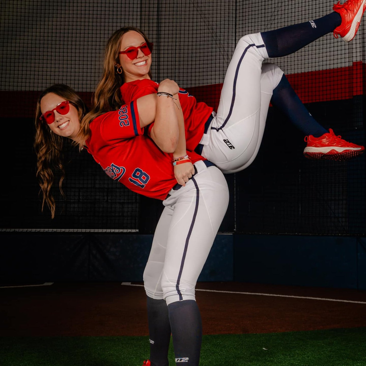 Two women in softball uniforms posing in a dugout.